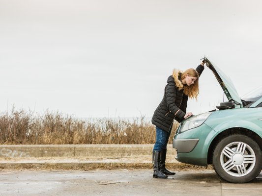 Girl standing over a broken down car looking under the hood.